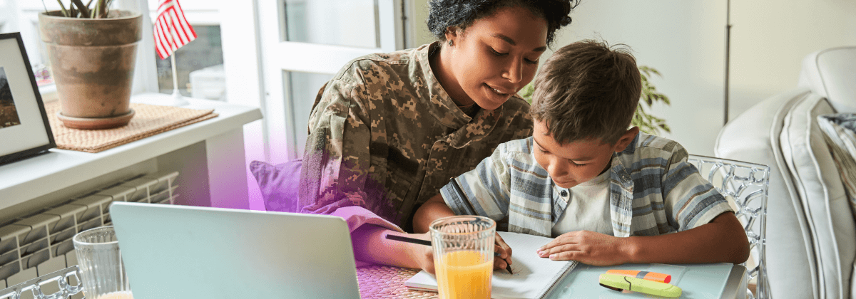 Mutter und Sohn sitzen an einem Tisch und zeichnen auf einem Blatt Papier, vor ihnen ein geöffneter Laptop. Die Mutter trägt eine Militäruniform.